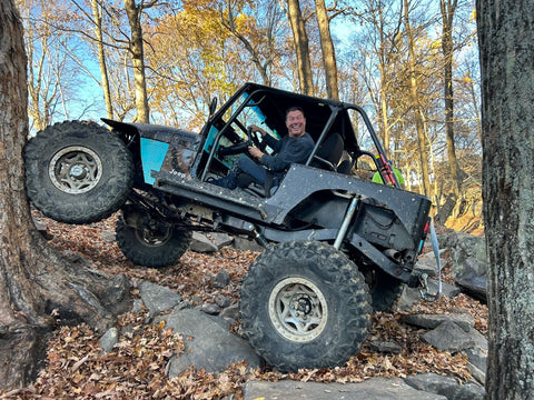 Eli smiling from a custom Jeep while climbing a rocky wooded trail
